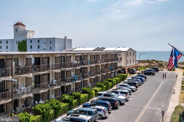 Three-story beachfront HOA with balconies overlooks a parking lot full of cars; an American flag, ocean, and sandy beach are visible in the background.