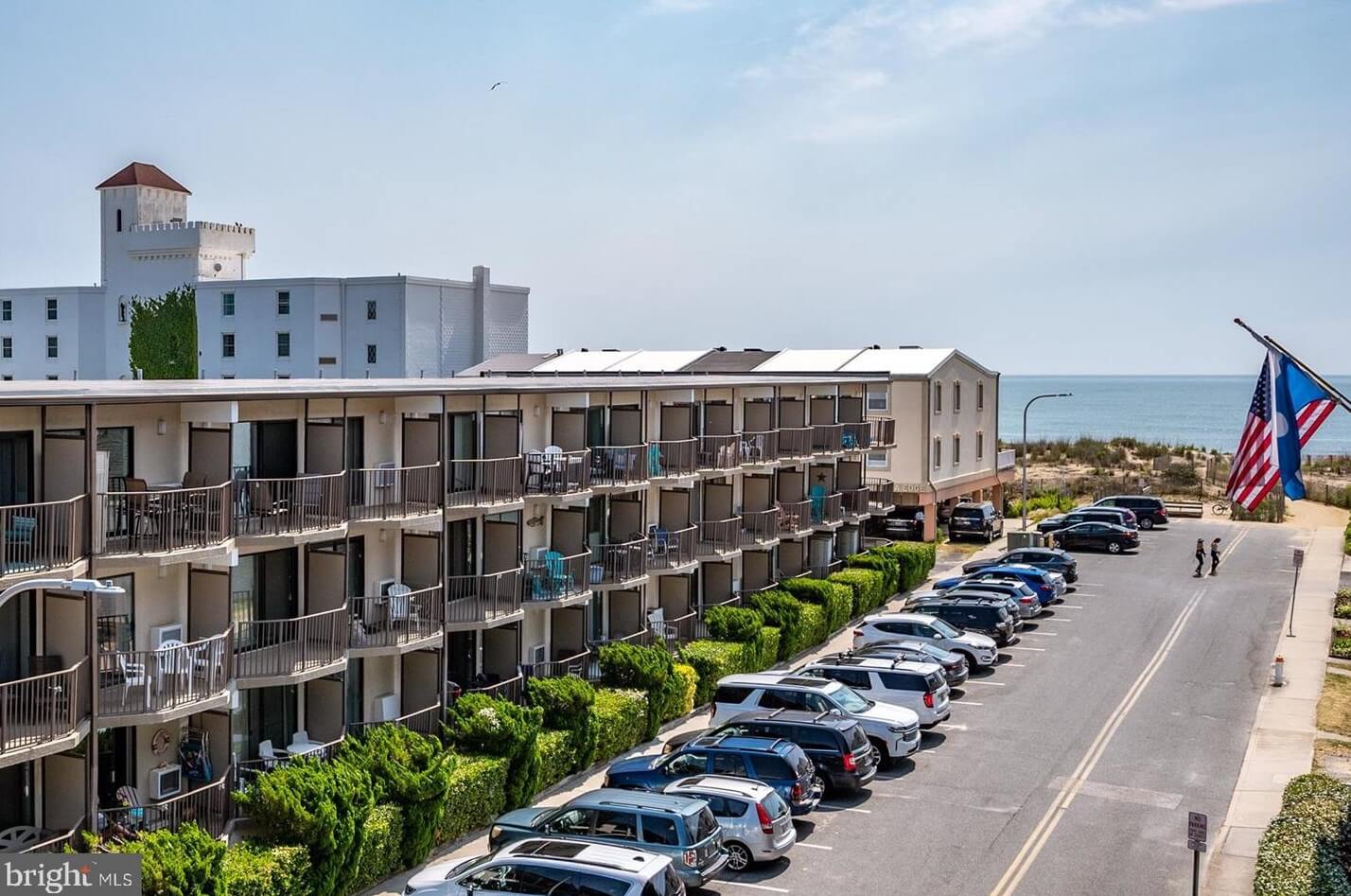 Three-story beachfront HOA with balconies overlooks a parking lot full of cars; an American flag, ocean, and sandy beach are visible in the background.